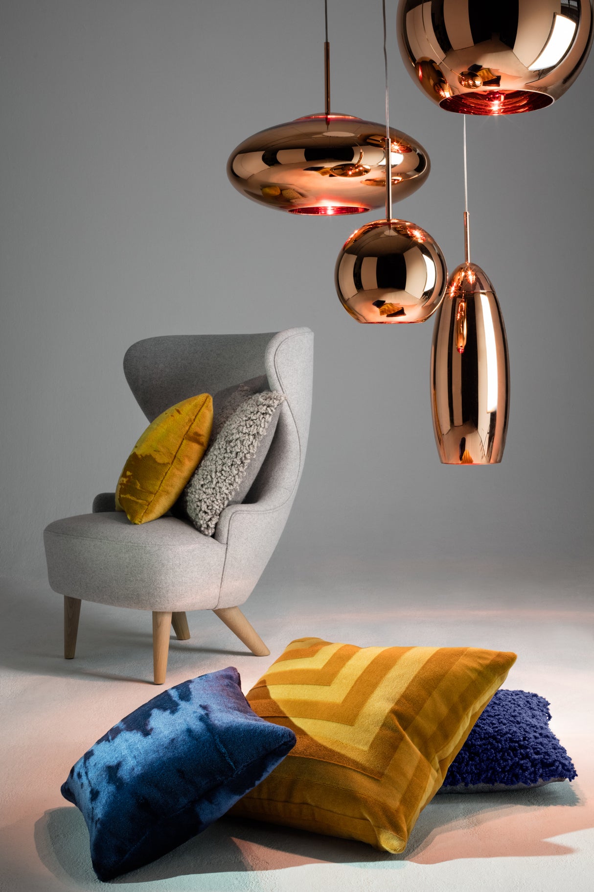 Gray wingback chair with mustard, gray, and textured cushions beside blue and mustard pillows under copper pendant lights
