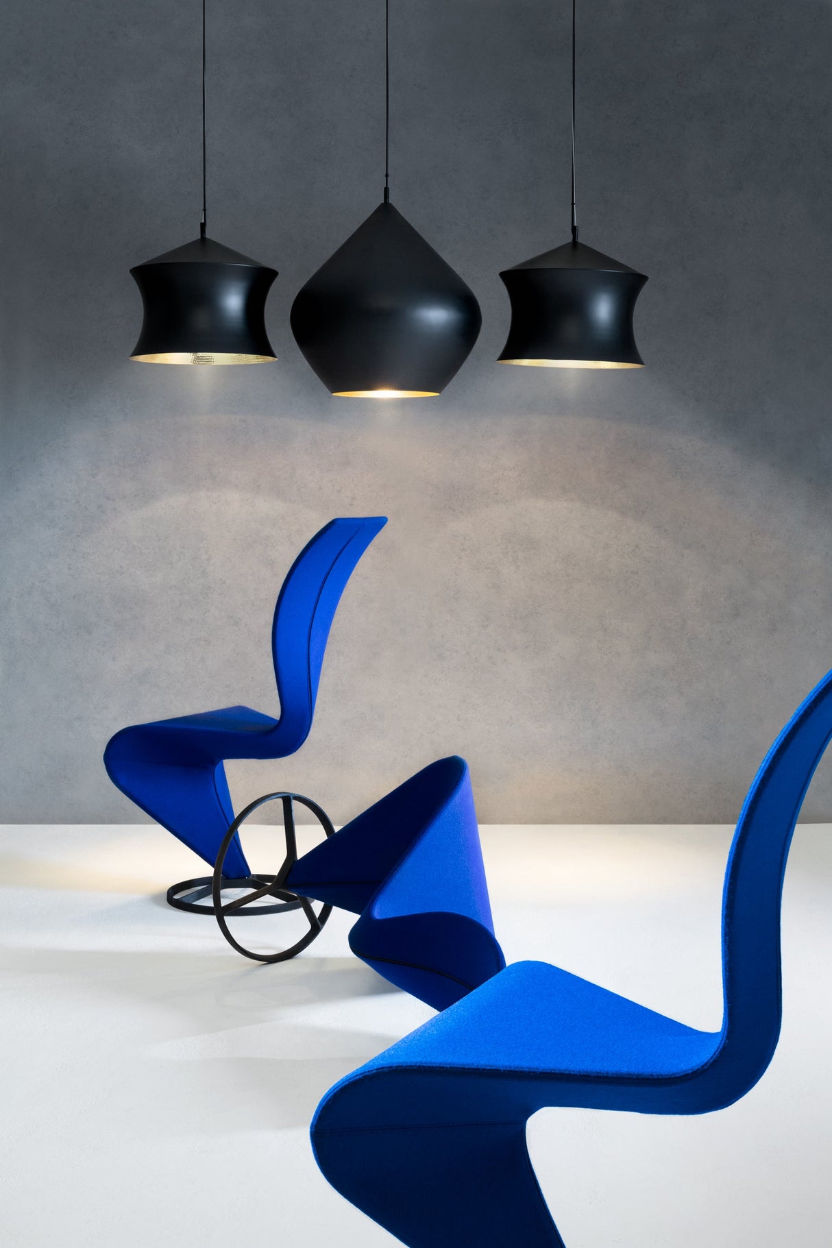 Three modern blue sculptural chairs arranged on a white floor under three black pendant LED lights in a minimalist room