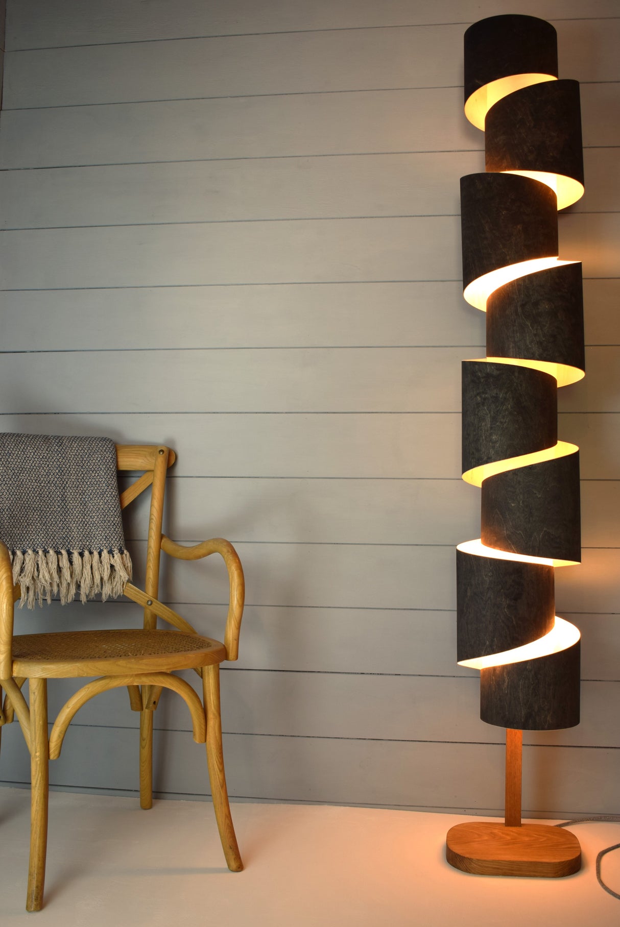 Modern spiraled wooden floor lamp with warm light near wooden chair with throw in cozy room