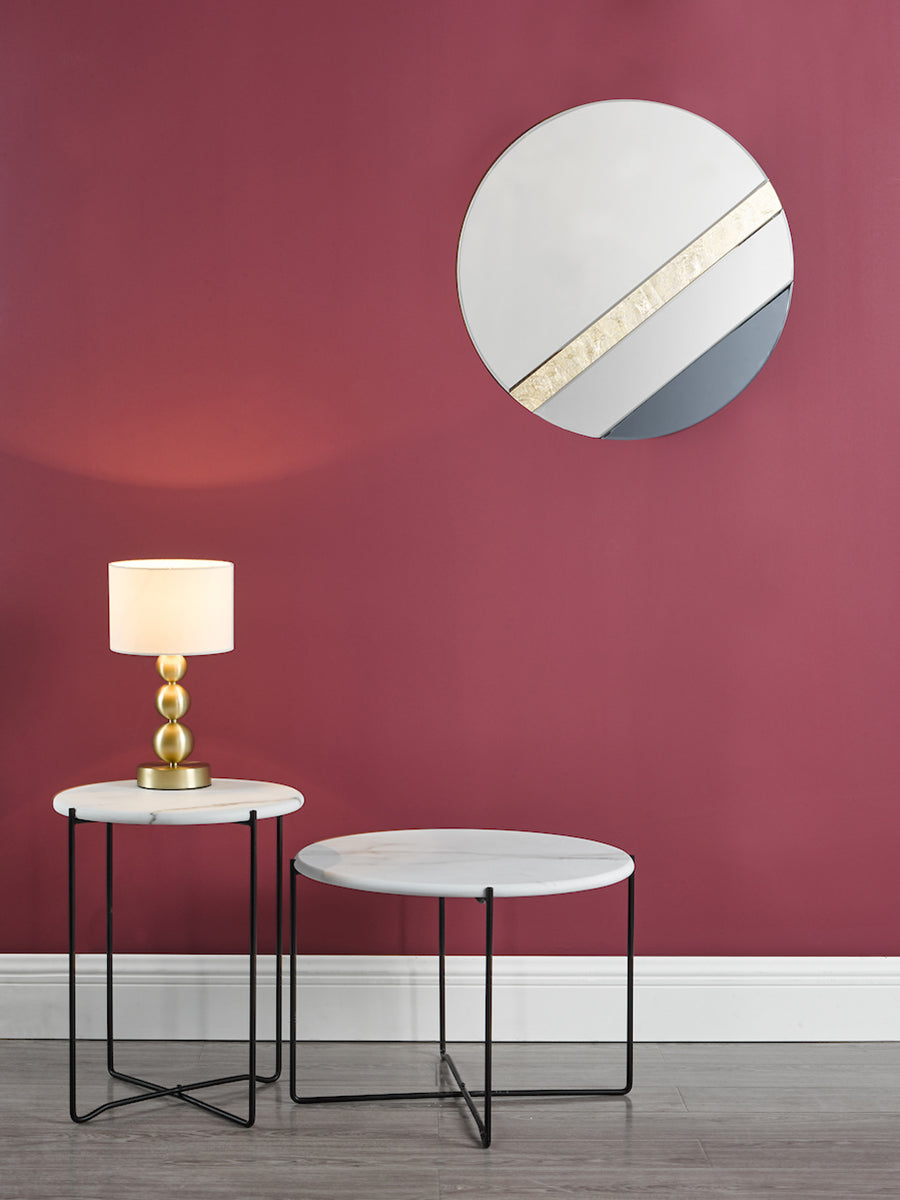 Modern round smoked mirror with gold leaf above marble side tables and gold lamp in room with burgundy wall