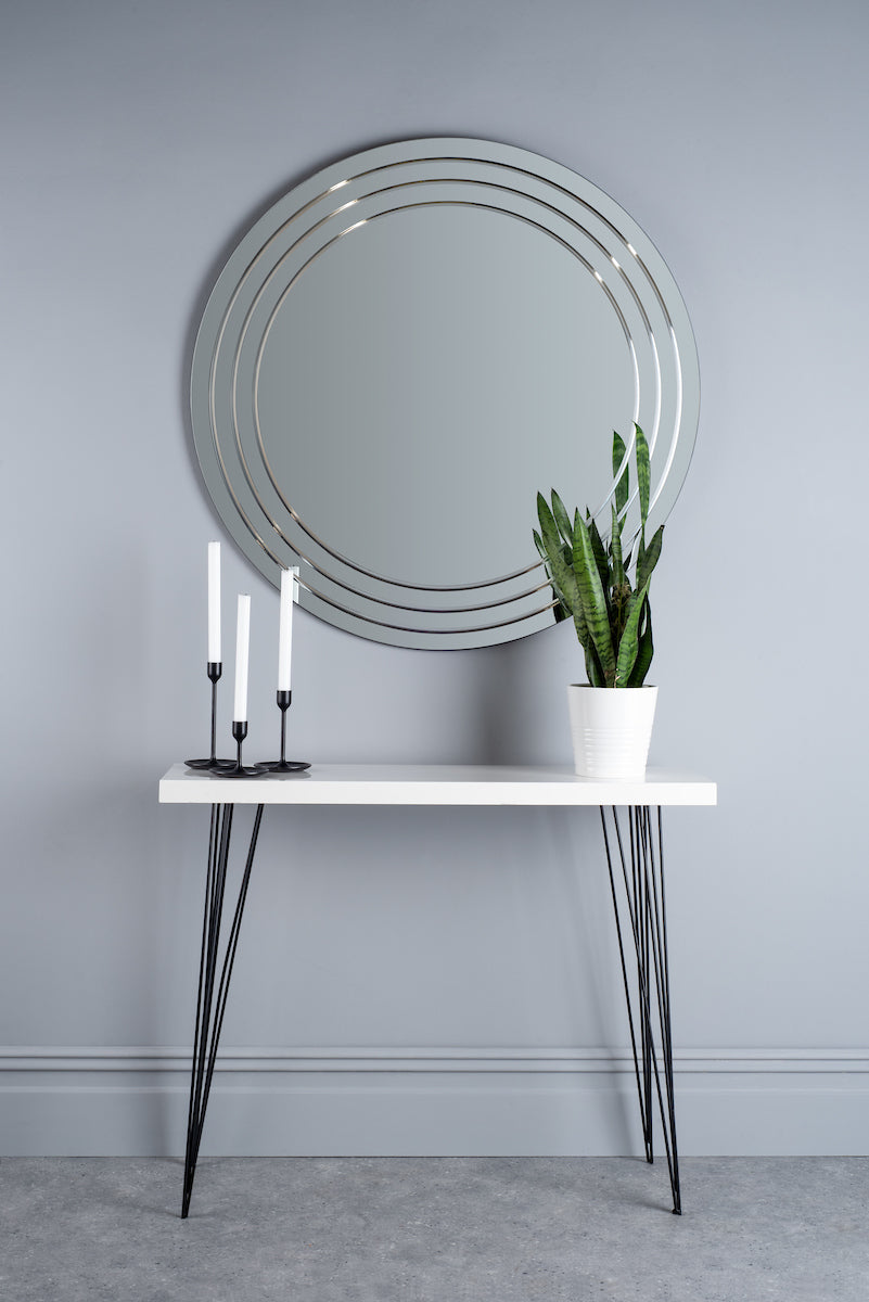Modern round bevelled mirror above white console table with black legs, candles, and potted snake plant