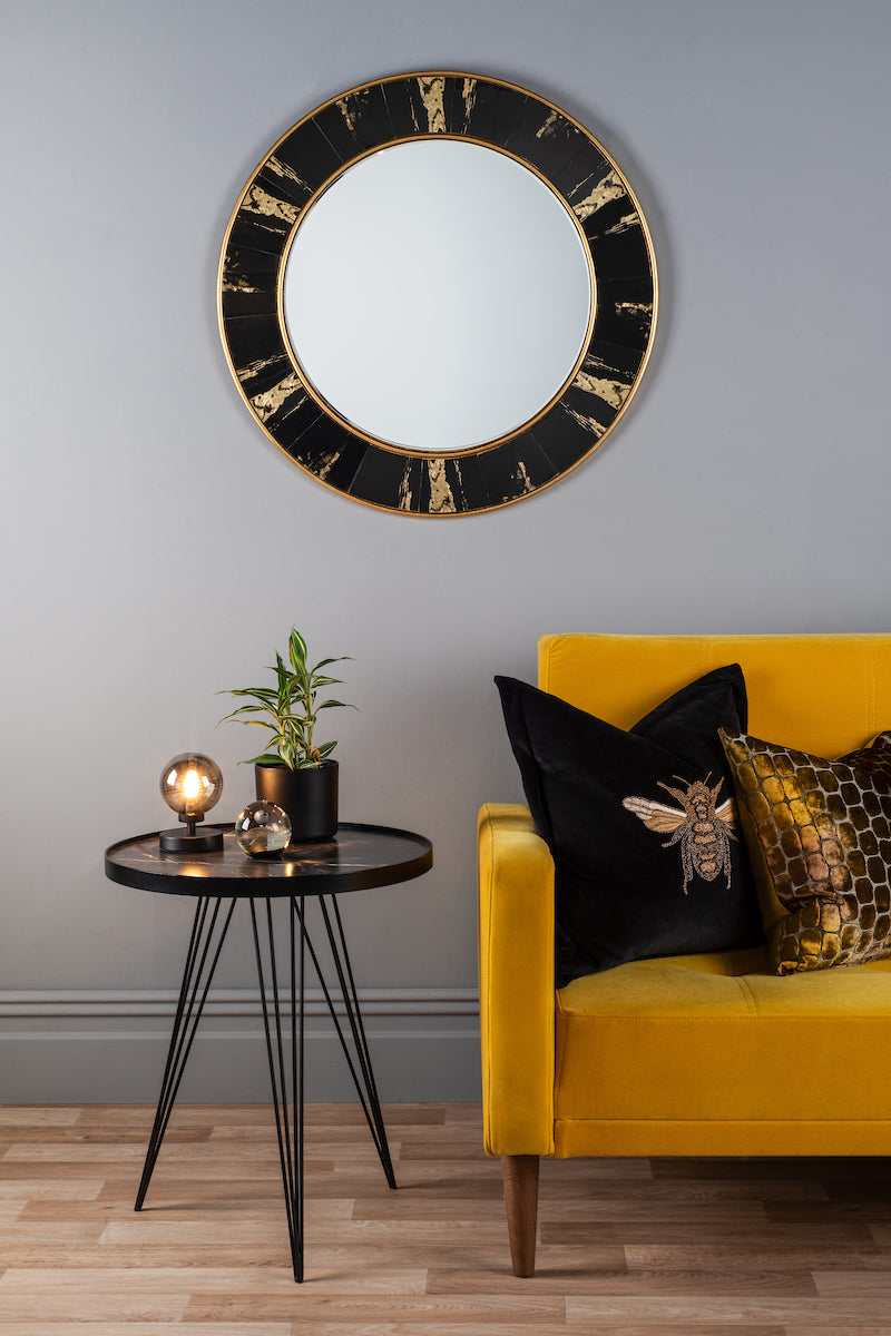 Round wall mirror with black and gold foil frame above black side table and yellow sofa with decorative pillows