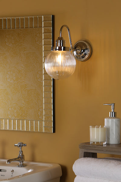 Chrome bathroom wall light with ribbed glass shade above white sink and towel on wooden shelf
