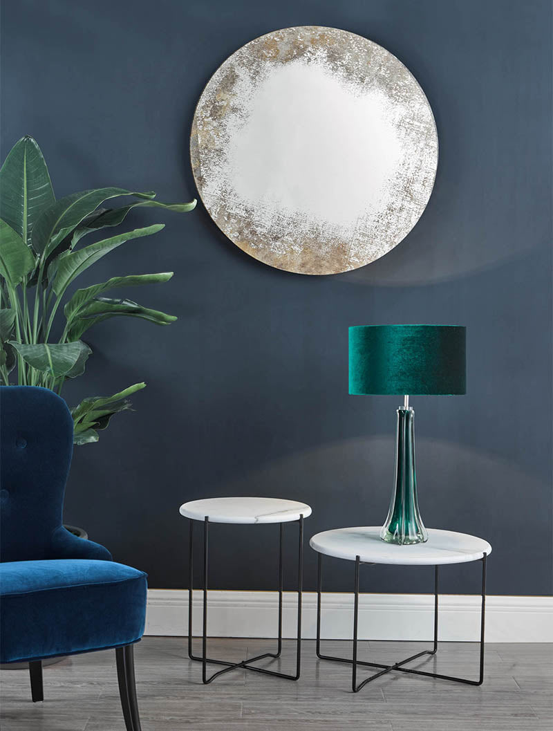 Round vintage mirror on dark blue wall above two white marble side tables, green velvet lamp, blue velvet chair, and large plant