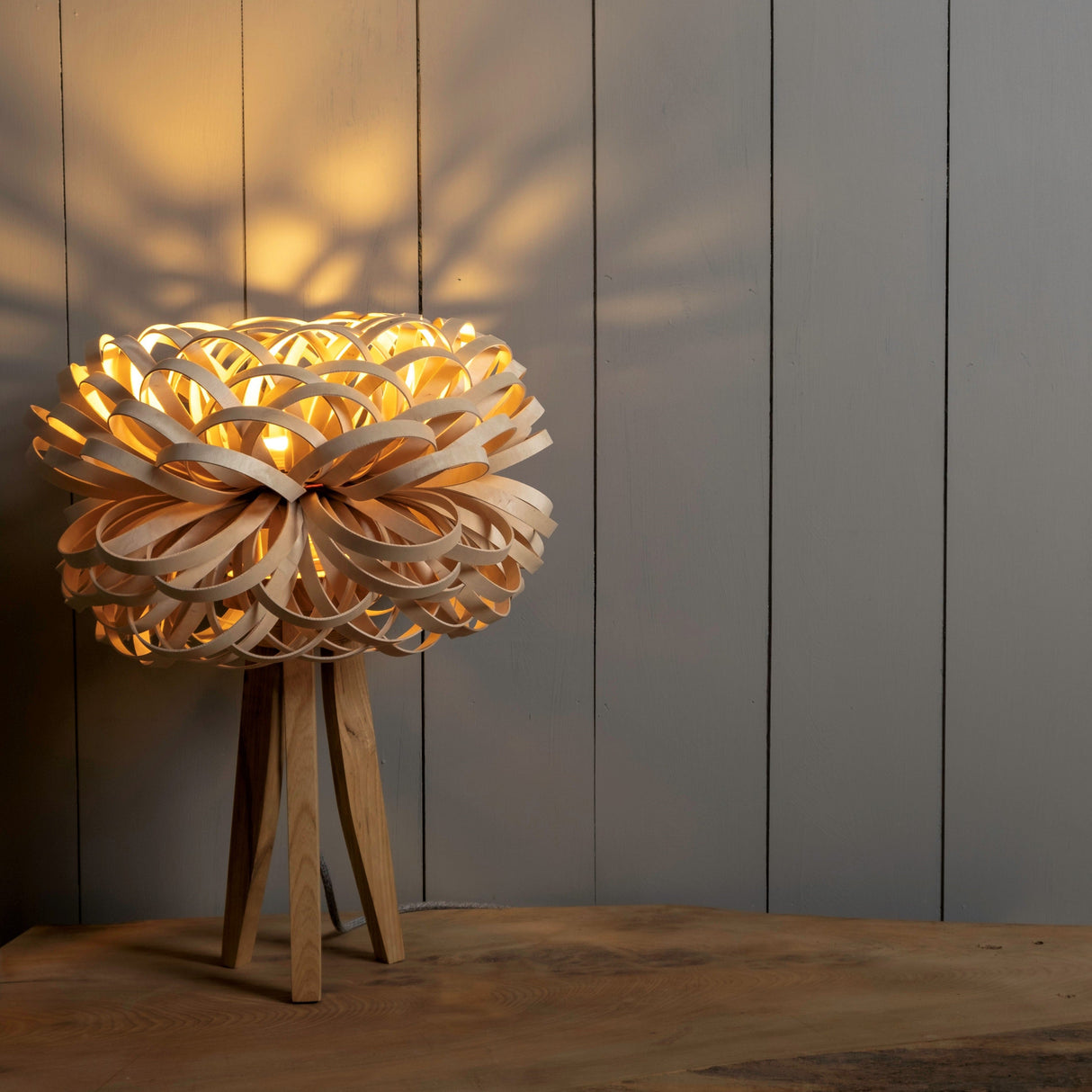 Wooden tripod table lamp with intricate allium-shaped shade emitting warm light on wooden surface