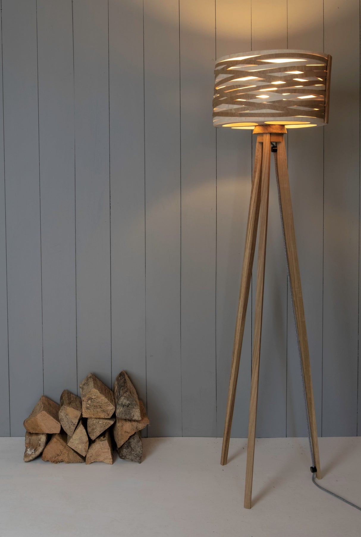 Modern wooden tripod floor lamp with unique cutout drum shade next to stacked firewood