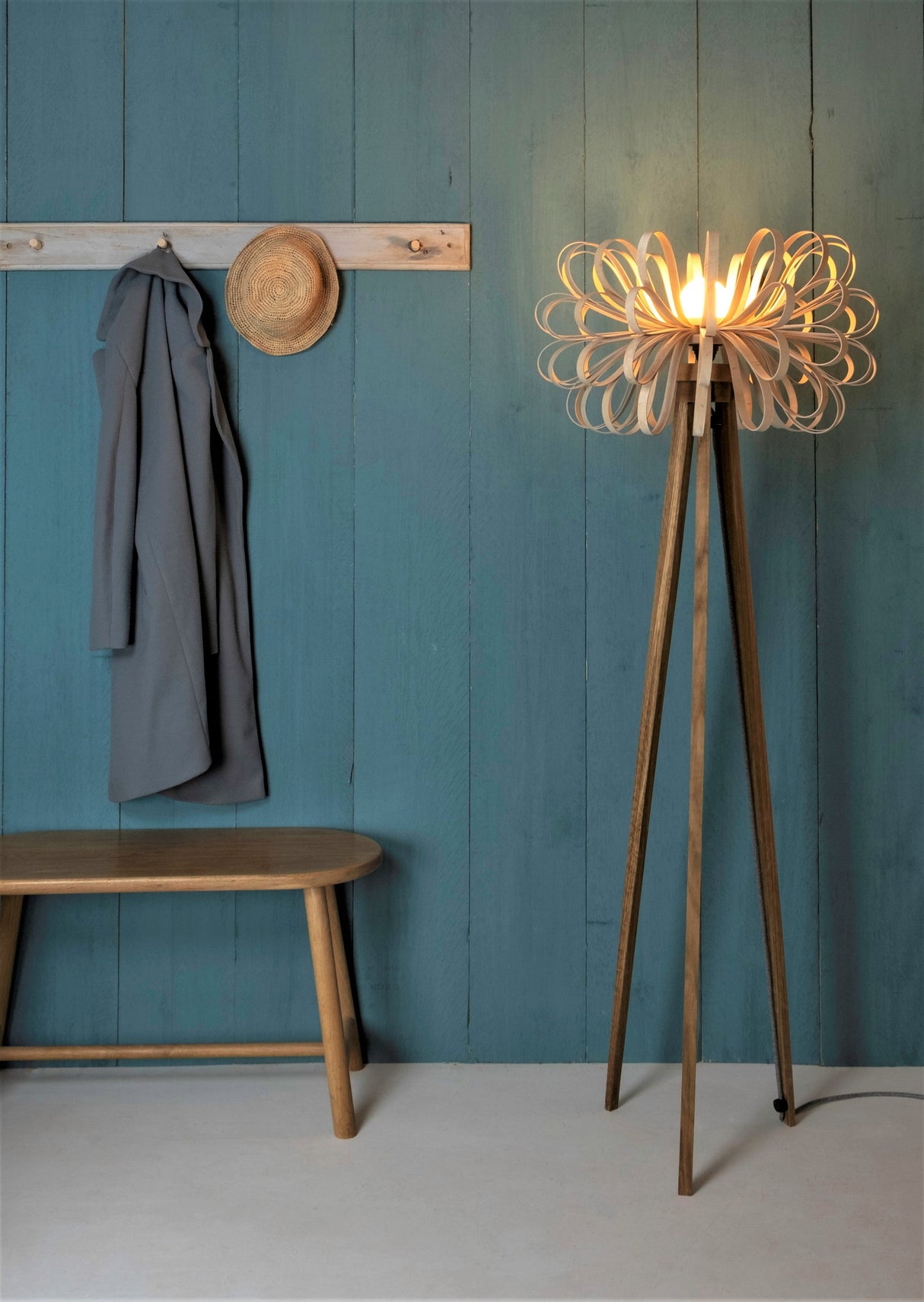 Modern wooden tripod floor lamp with artistic looped shade glowing beside a bench and coat on teal plank wall