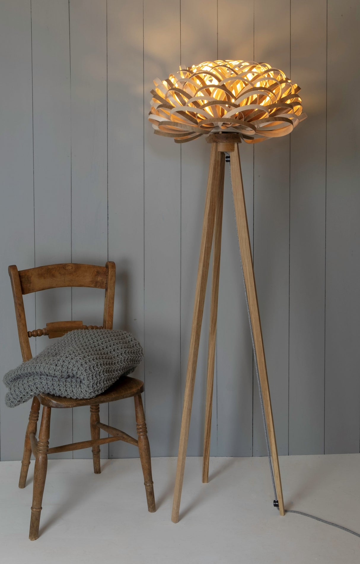 Wooden tripod floor lamp with intricate circular shade next to rustic wooden chair with gray knitted blanket