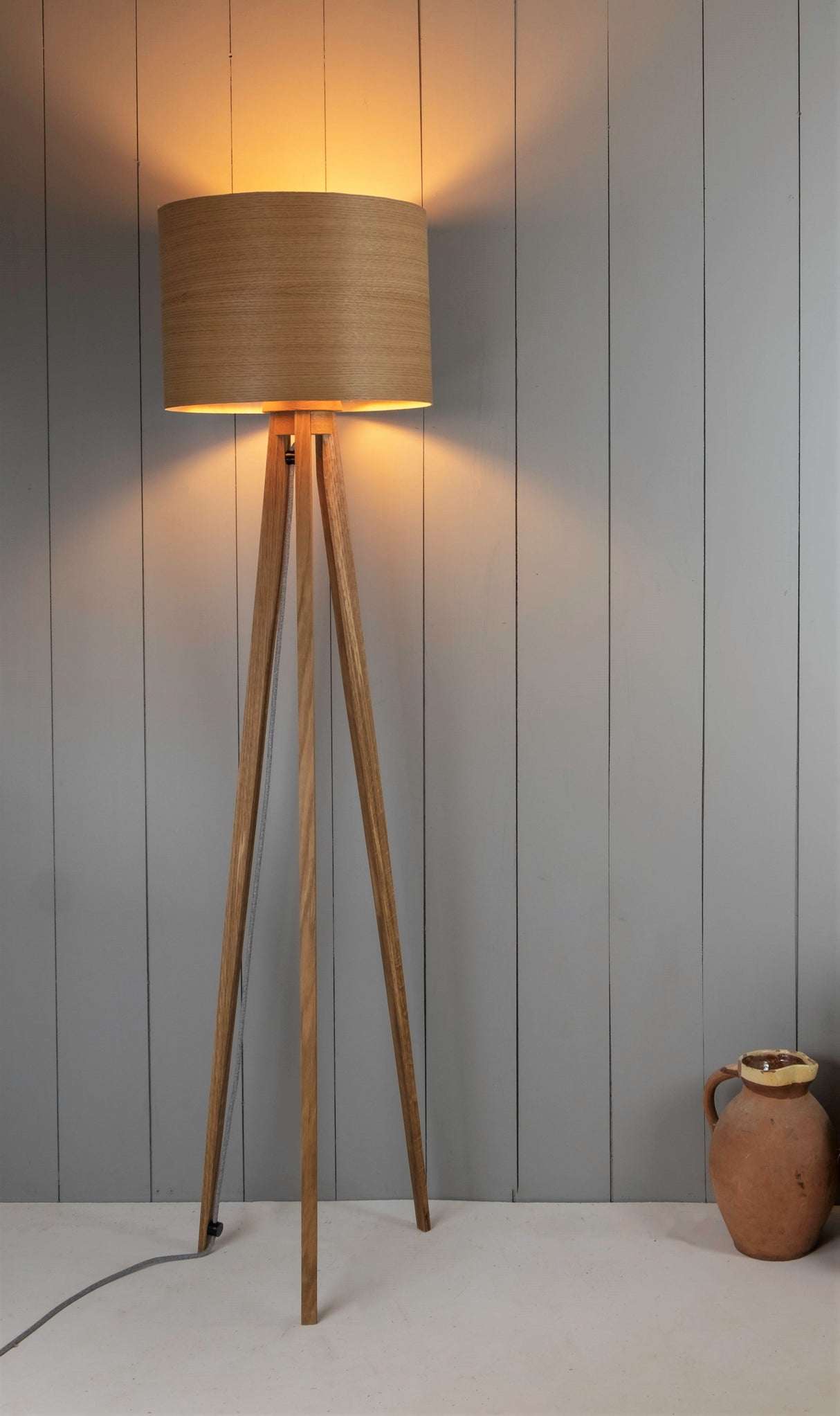 Tripod wooden floor lamp with cylindrical shade illuminated against gray paneled wall, ceramic jug on floor