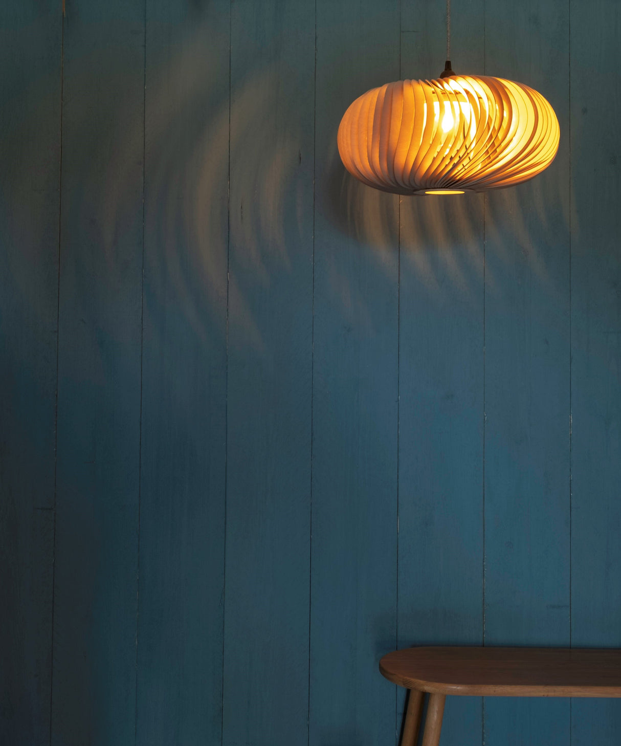 Modern wooden pendant ceiling light glowing over wooden bench against teal panel wall