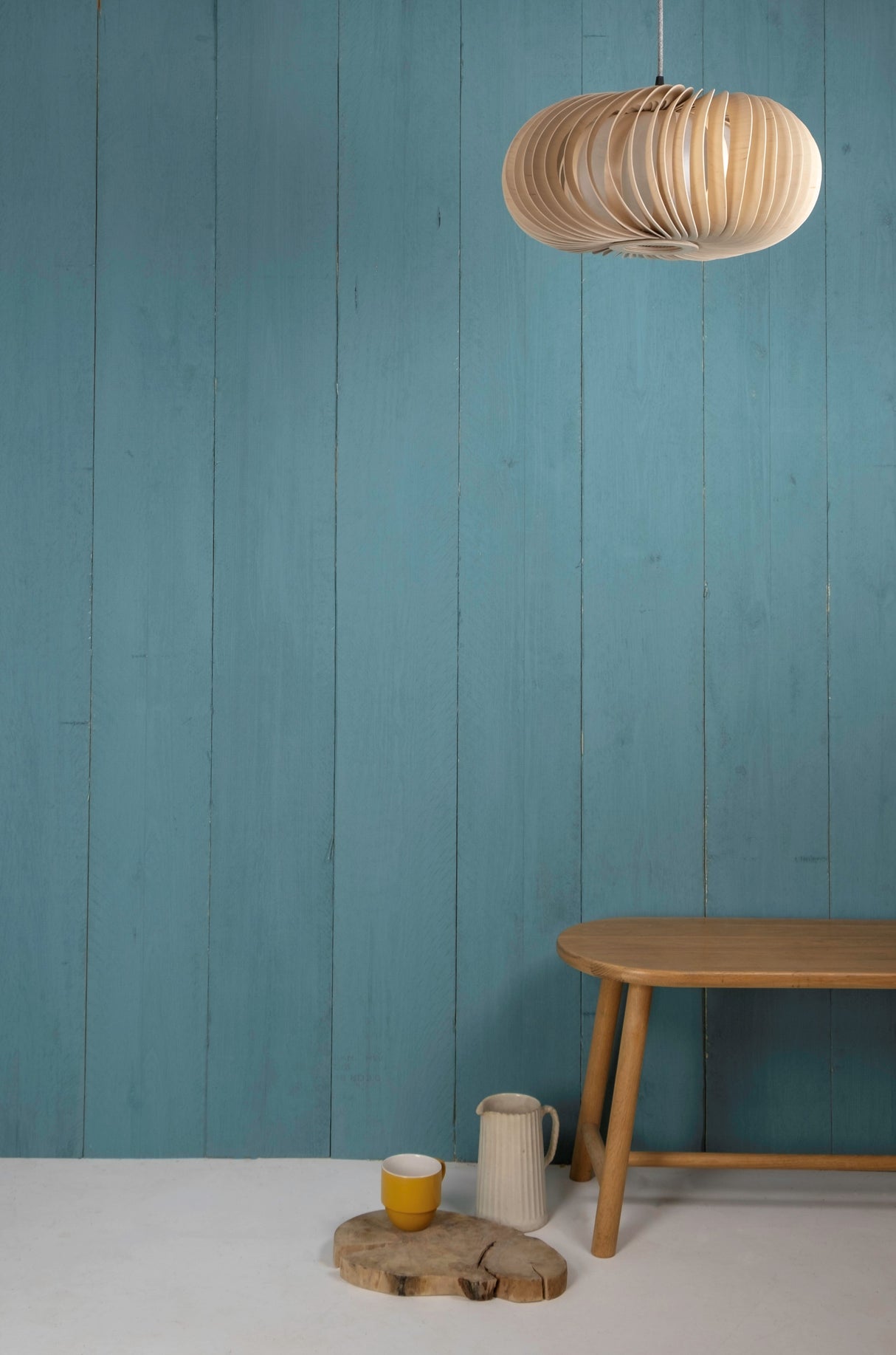 Modern wooden pendant light above wooden bench with rustic cup and pitcher against blue paneled wall