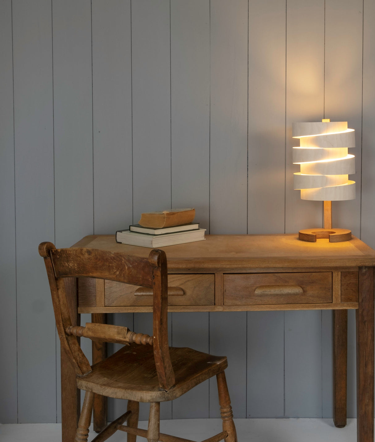 Wooden desk and chair with vintage books and modern spiral design table lamp against gray paneled wall