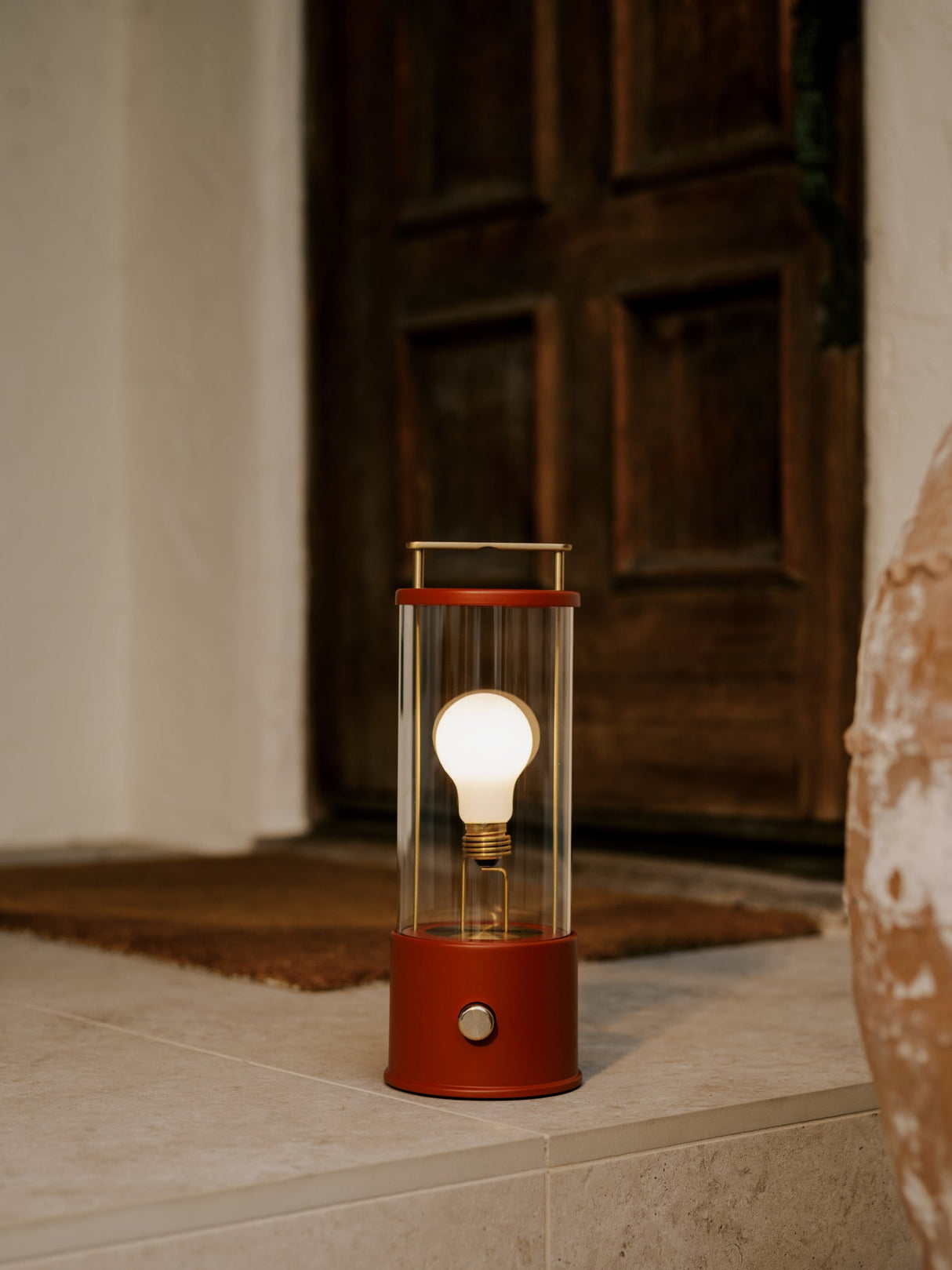 Tala Muse portable lamp in Pomona Red with glowing bulb on indoor tiled floor near wooden door