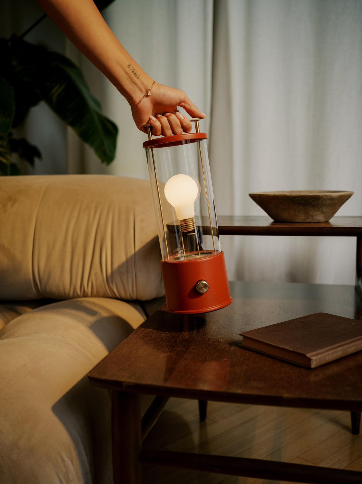 Hand holding a red portable lamp with a glowing bulb next to beige sofa and wooden table