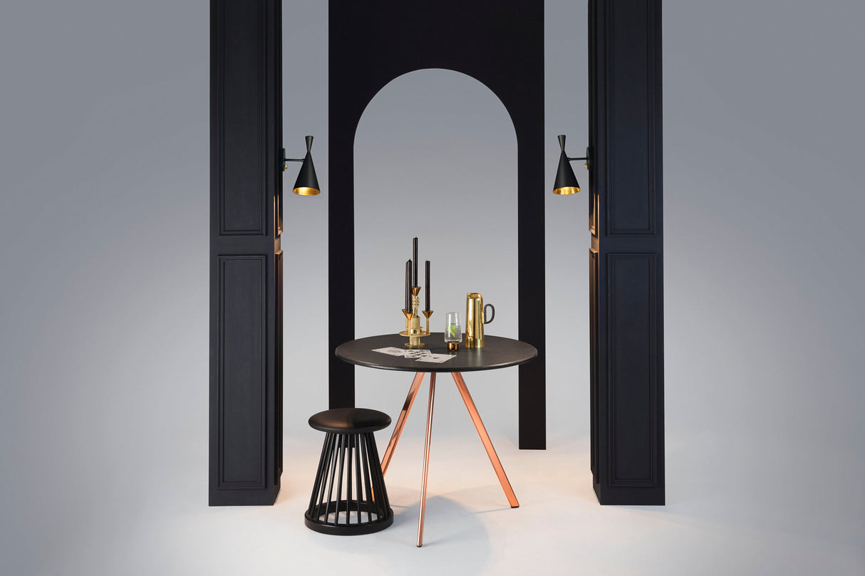 Modern black and copper round table with black stool, brass candlesticks, pitcher, and wall lights in minimalist setting