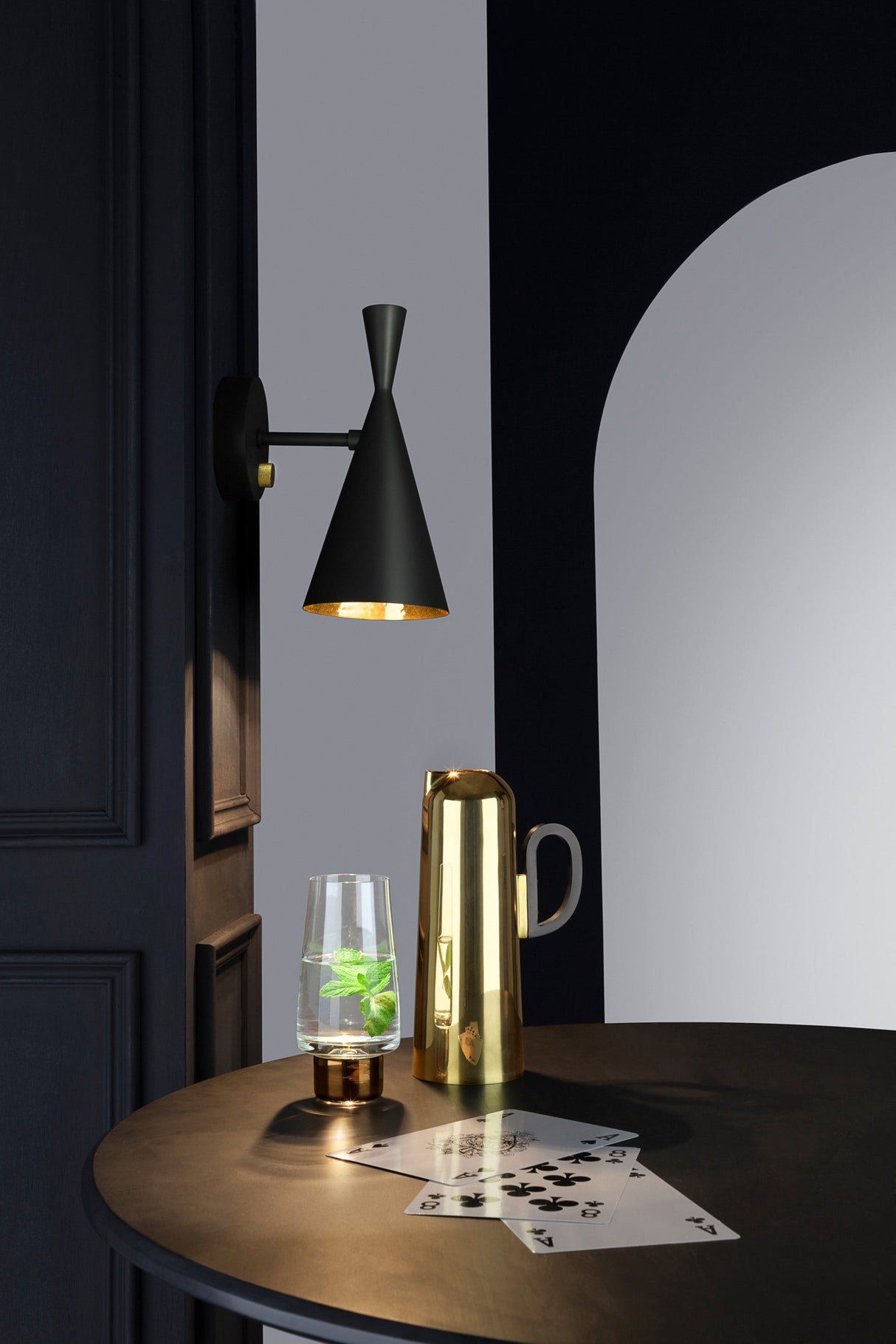 Tom Dixon Beat black wall light with brass accents above glass with mint and gold pitcher on black round table with playing cards