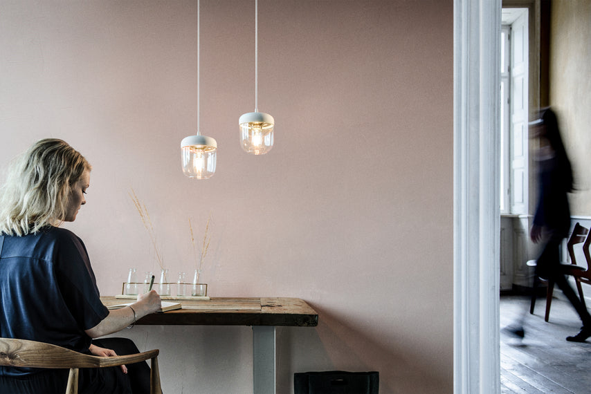 Woman writing at wooden table under two modern white polished steel pendant lights in minimalist room