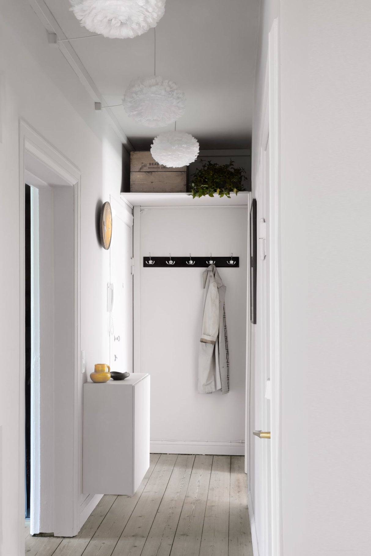 Minimalist white hallway with feather pendant lights, floating cabinet, wooden floor, coat rack with beige jacket