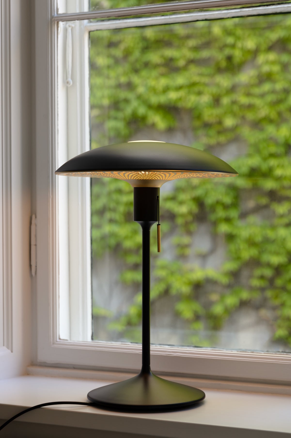 Umage Manta Ray black brass table lamp on a white windowsill with green foliage outside