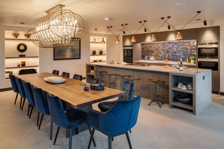 Modern dining room with wooden table, blue velvet chairs, chandelier, kitchen island, and copper pendant lights