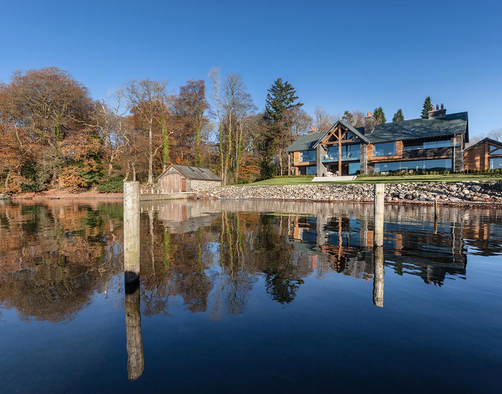 Modern lakeside house with large windows and wooden beams reflecting on calm water under clear blue sky