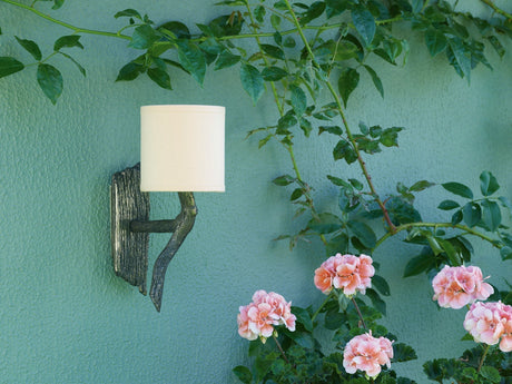 Outdoor wall sconce with white lampshade on green textured wall surrounded by green leaves and pink flowers