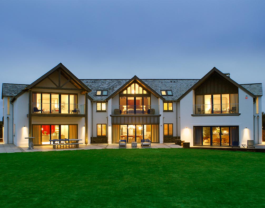 Modern large two-story house with illuminated windows, balconies, and green lawn at dusk
