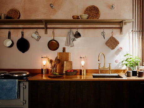 Cozy kitchen countertop with brass faucet, hanging pans, wooden utensils, soft lighting, and potted plants