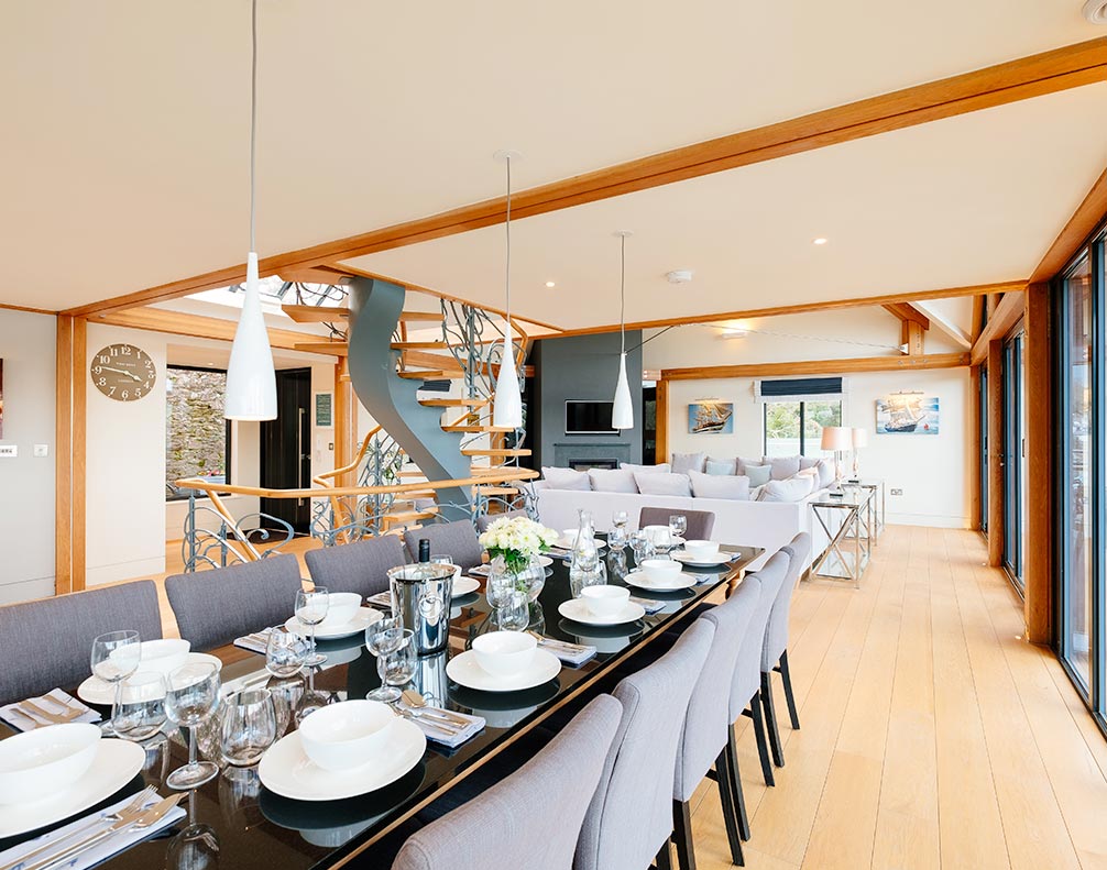 Modern open dining and living room with large glass table, gray chairs, spiral staircase, and wooden flooring