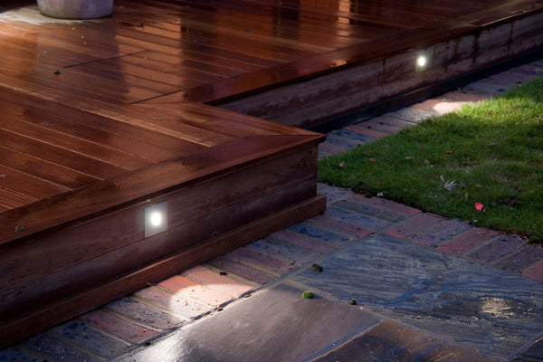 Outdoor wooden deck at night with built-in LED step lights illuminating brick and stone patio, green lawn