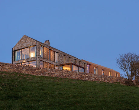 Modern stone and wood house on hill with large windows at dusk and clear blue sky