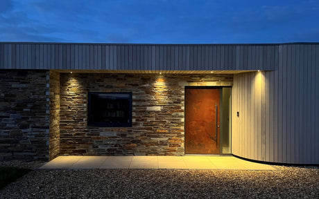 Modern house entrance at dusk with stone wall, rust-colored door, window, and warm outdoor lighting