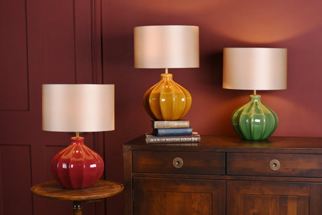Three ceramic table lamps in red, mustard, and green on wooden furniture against dark red wall