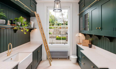 Heritage kitchen with dark green cabinets, brass fixtures, white countertops, and a ladder by a large window