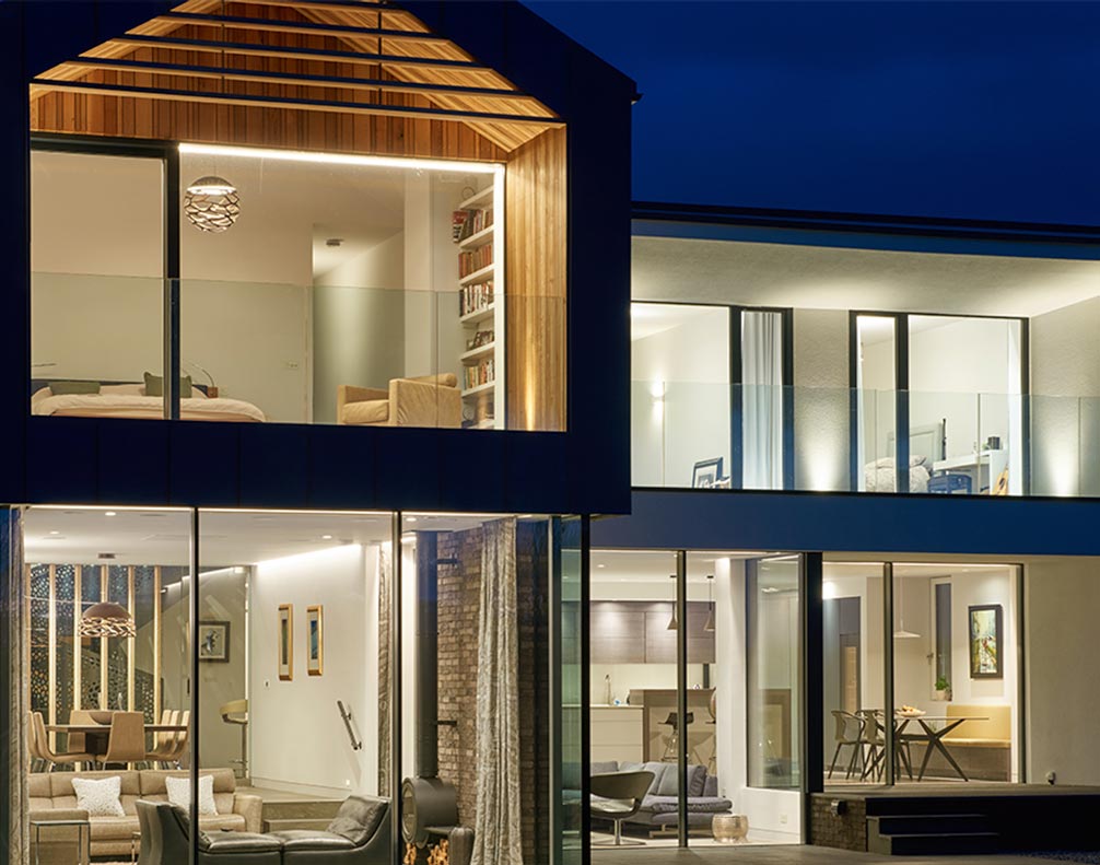 Modern two-story house with large glass windows, illuminated interior at night, showing living and dining areas