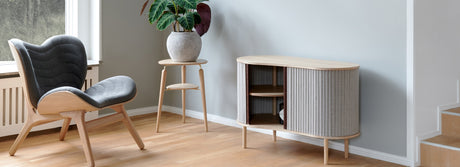 Modern living room with wooden lounge chair, small plant table, and light wood sideboard with fabric doors