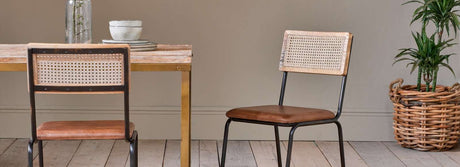 Wood dining table with metal legs, two brown leather and cane back chairs, ceramic bowl set, and potted plant in wicker basket