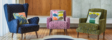 Dark green velvet sofa with patterned cushions, wooden wall, potted plant, and geometric curtain