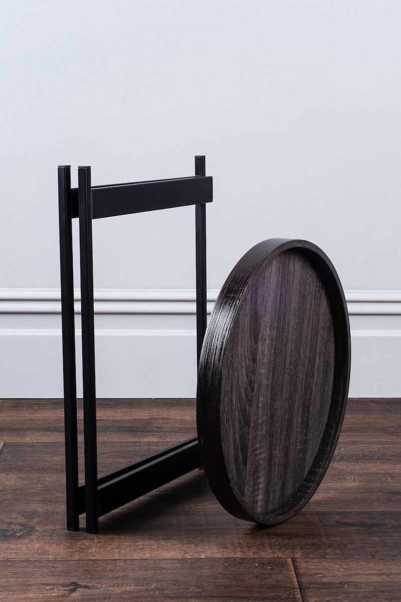 Round black wooden tray with black metal folding stand on wooden floor against white wall