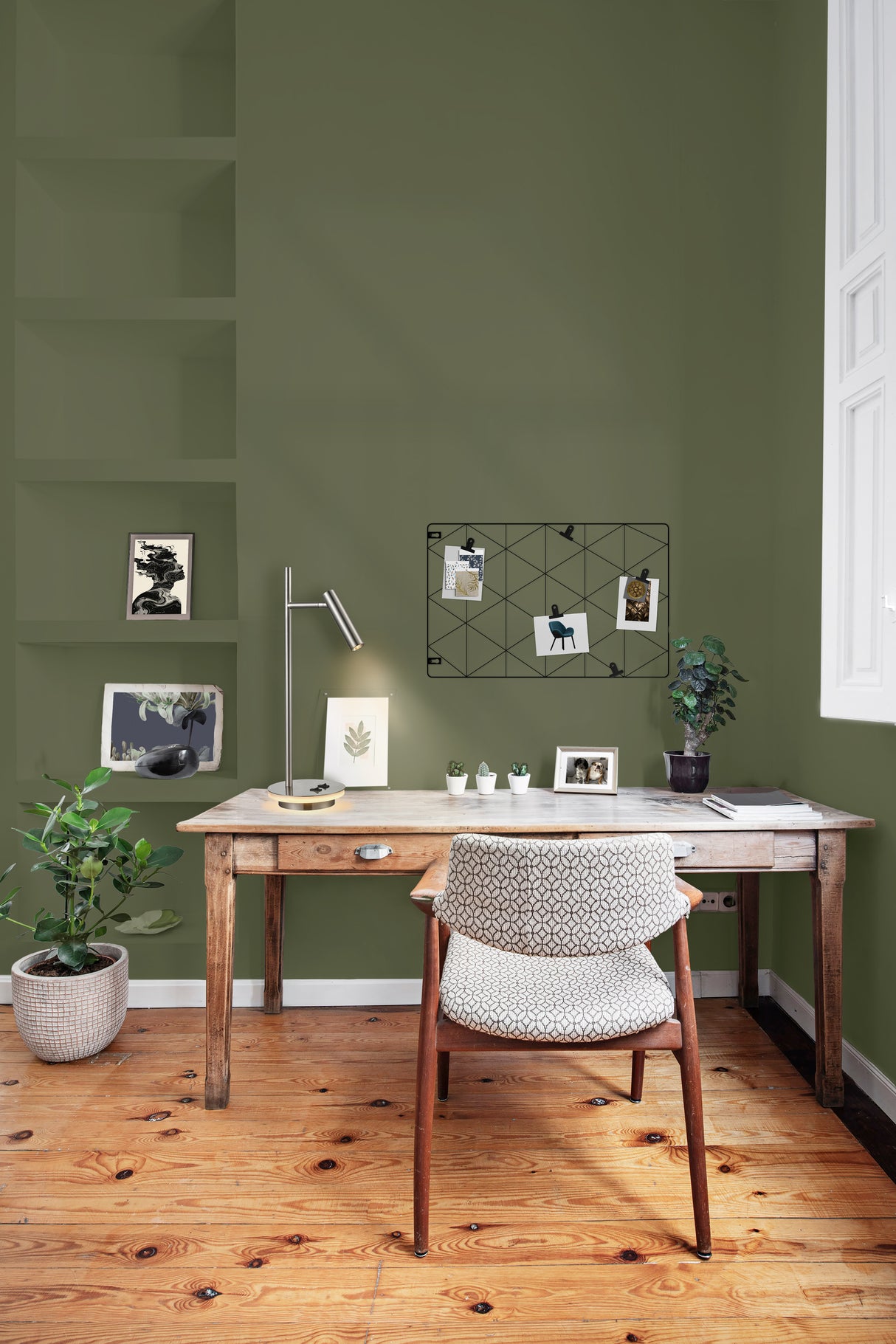 Cozy home office with wooden desk, patterned chair, green walls, potted plants, and desk lamp