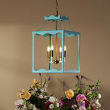 A turquoise blue hanging lantern-style chandelier with lit candle bulbs is suspended above a round table decorated with a large floral arrangement in pink, white, yellow, and burgundy flowers against a warm beige wall.