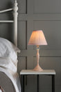 Soft glowing pink bedside lamp on white table next to white metal bed frame and pillows