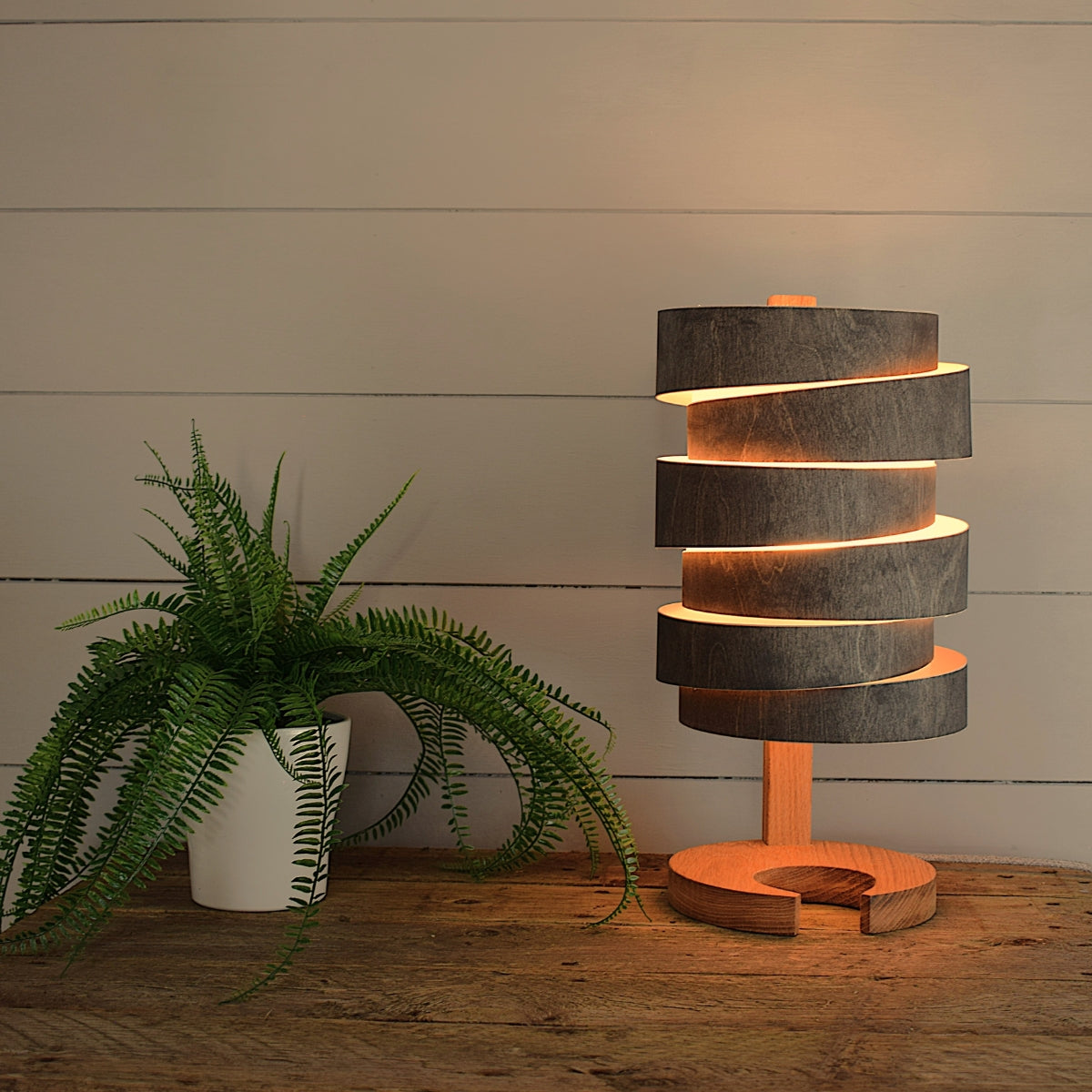 Modern wooden wrap table lamp emitting warm light on rustic wooden surface next to green potted fern