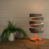 Modern wooden wrap table lamp emitting warm light on rustic wooden surface next to green potted fern